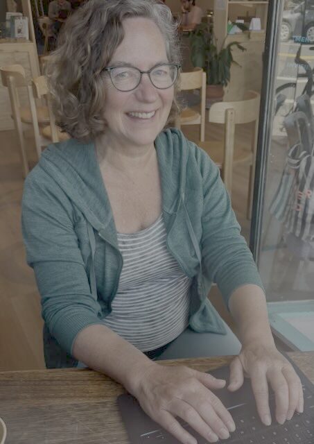 Cate, woman at a computer at a desk, smiling at the camera, wearing green glasses, a green jacket, and a striped tshirt